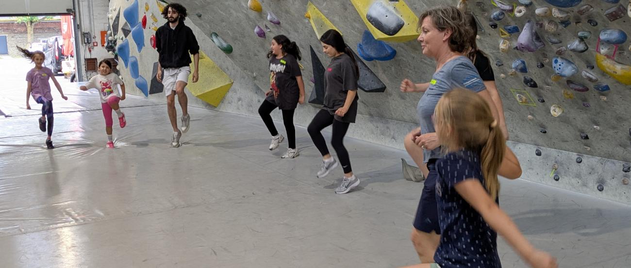 Kinder springen im Halbkreis als Aufwärmübung vor einer Kletterwand.