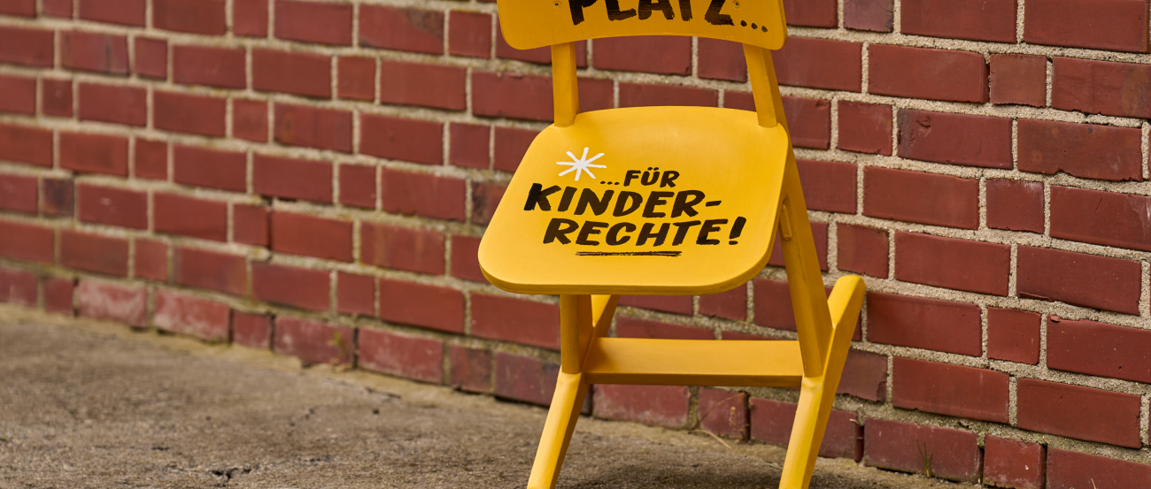 Ein gelber Stuhl lehnt an einer Hauswand. Auf dem Stuhl findet sich die Aufschrift 'Ein Platz...' und 'für Kinderrechte'.