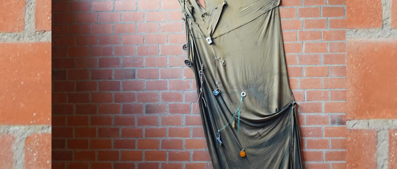 Dunkler Stoff vor Backsteinwand, mit Objekten behangen, formt die Kontur einer Knienden