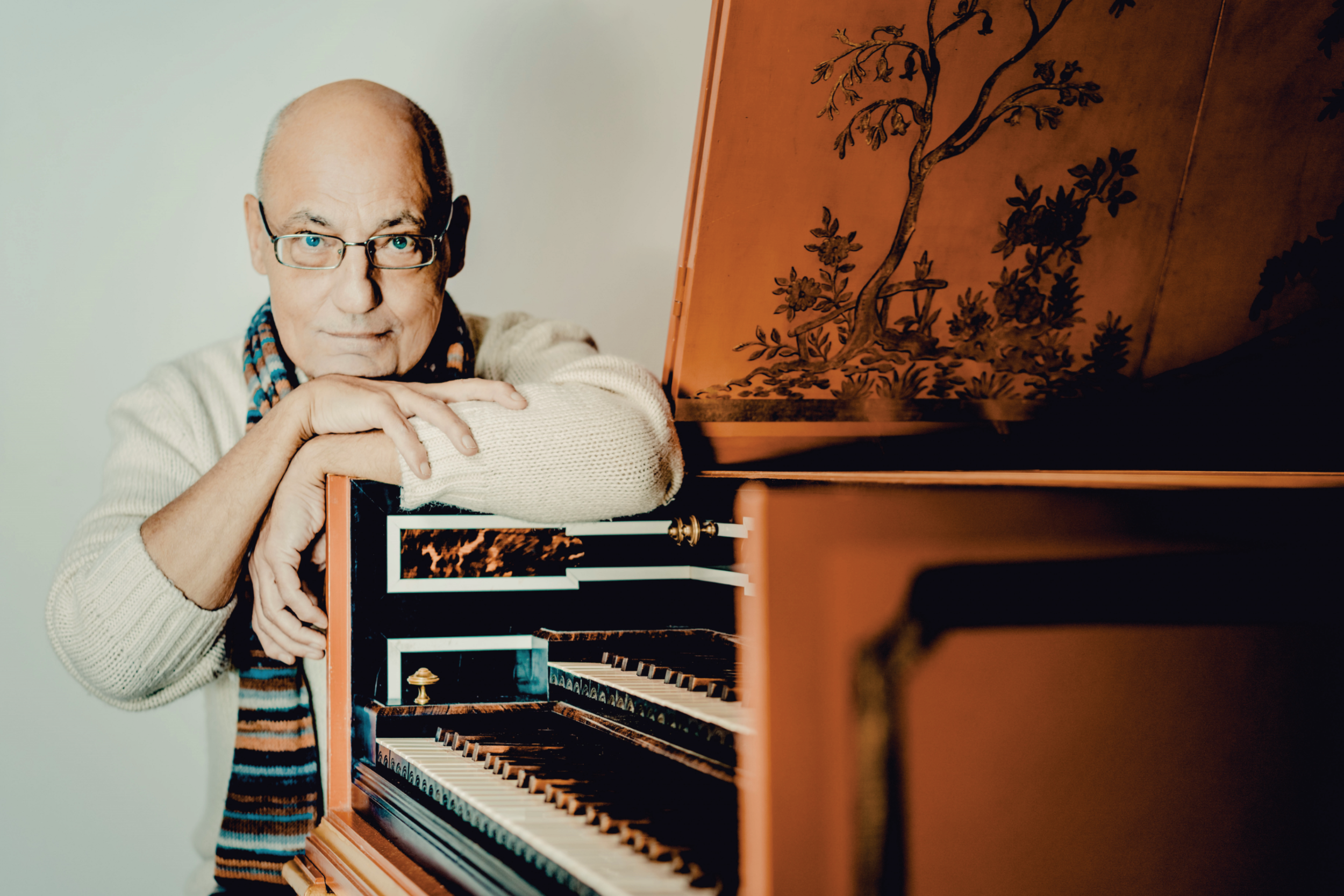 Älterer Mann mit Brille und Schal lehnt an einem historischen Tasteninstrument.