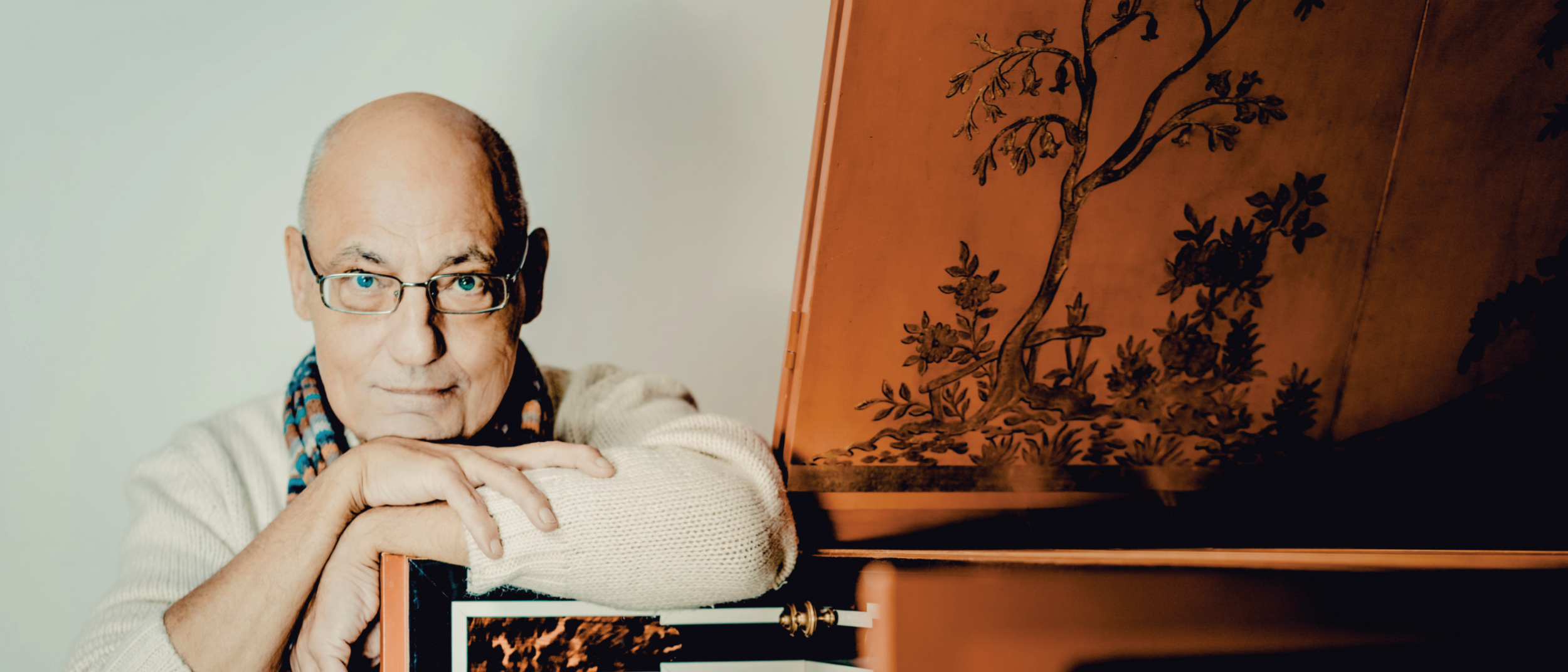 Älterer Mann mit Brille und Schal lehnt an einem historischen Tasteninstrument.