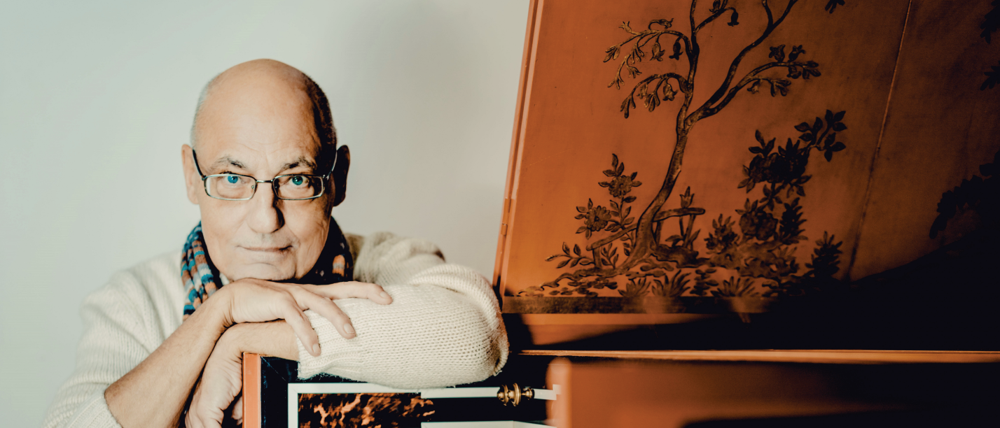 Älterer Mann mit Brille und Schal lehnt an einem historischen Tasteninstrument.