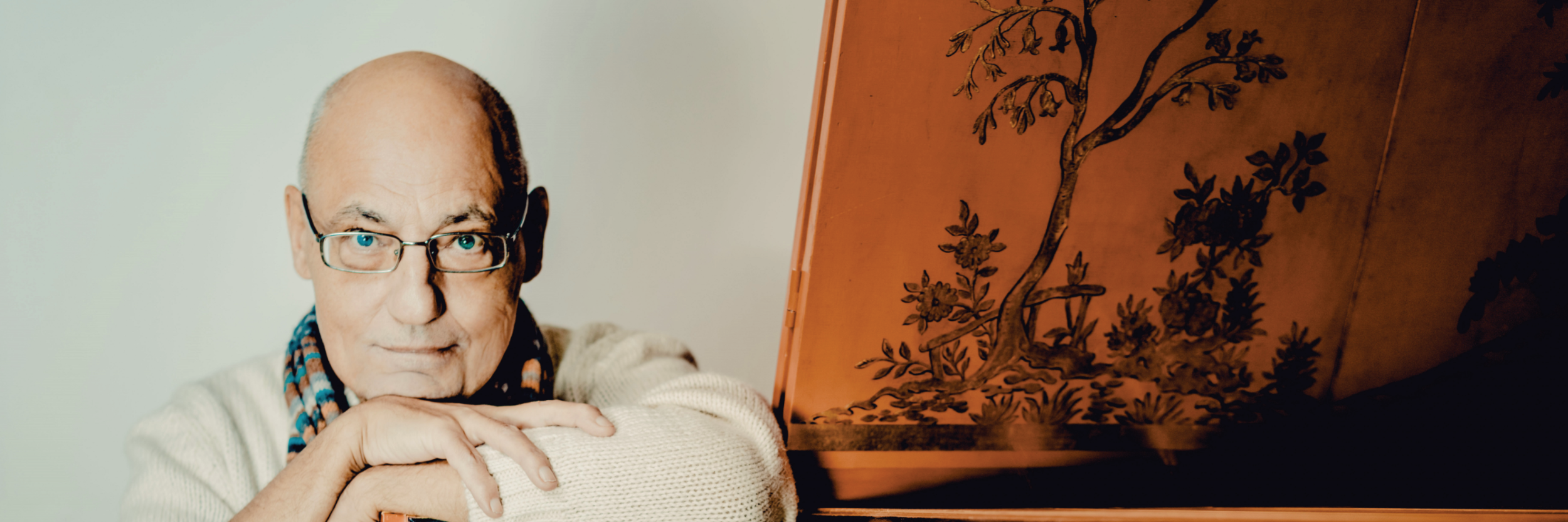 Älterer Mann mit Brille und Schal lehnt an einem historischen Tasteninstrument.