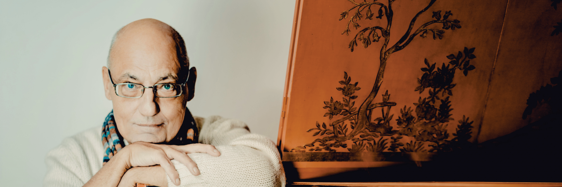 Älterer Mann mit Brille und Schal lehnt an einem historischen Tasteninstrument.
