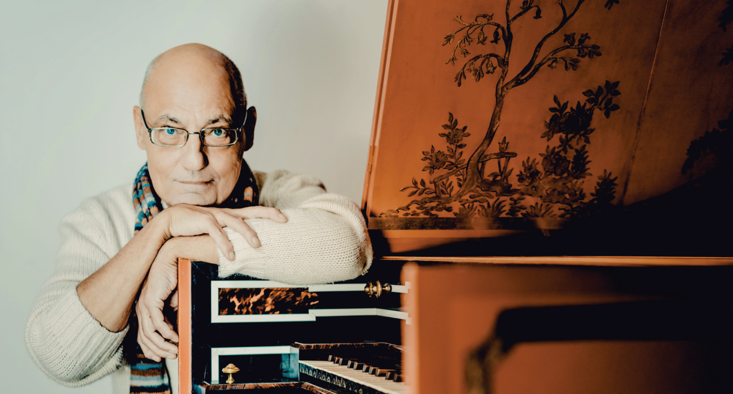 Älterer Mann mit Brille und Schal lehnt an einem historischen Tasteninstrument.