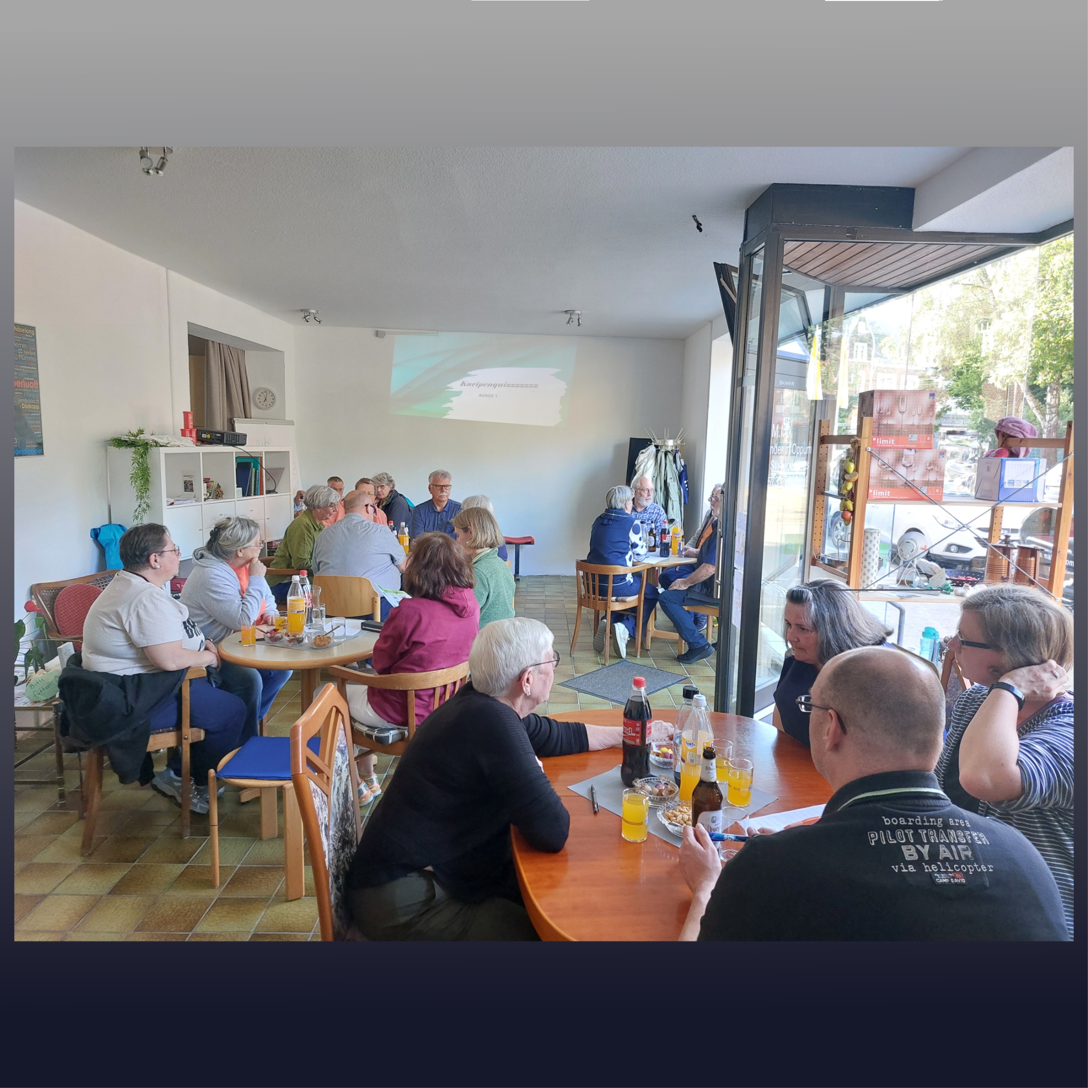 Menschen sitzen in Gruppen um Tische und schauen zu einem Bild an der Wand.