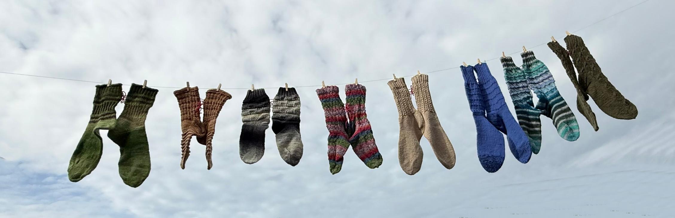 Mehrere bunte gestrickte Socken hängen an einer Wäscheleine vor bewölktem Himmel.