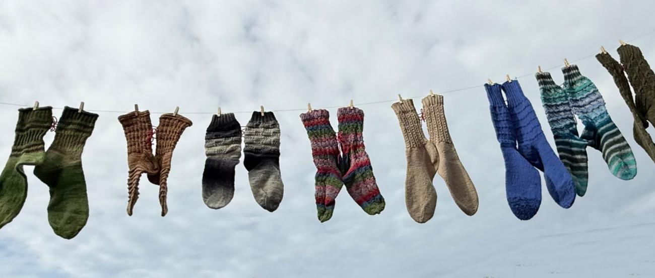 Mehrere bunte gestrickte Socken hängen an einer Wäscheleine vor bewölktem Himmel.