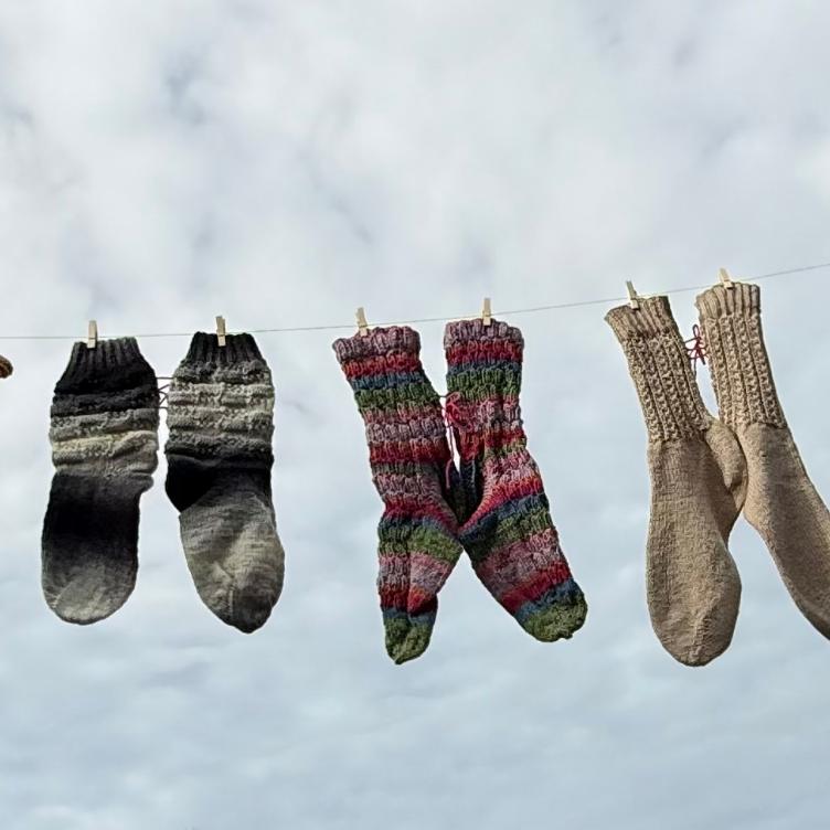 Mehrere bunte gestrickte Socken hängen an einer Wäscheleine vor bewölktem Himmel.