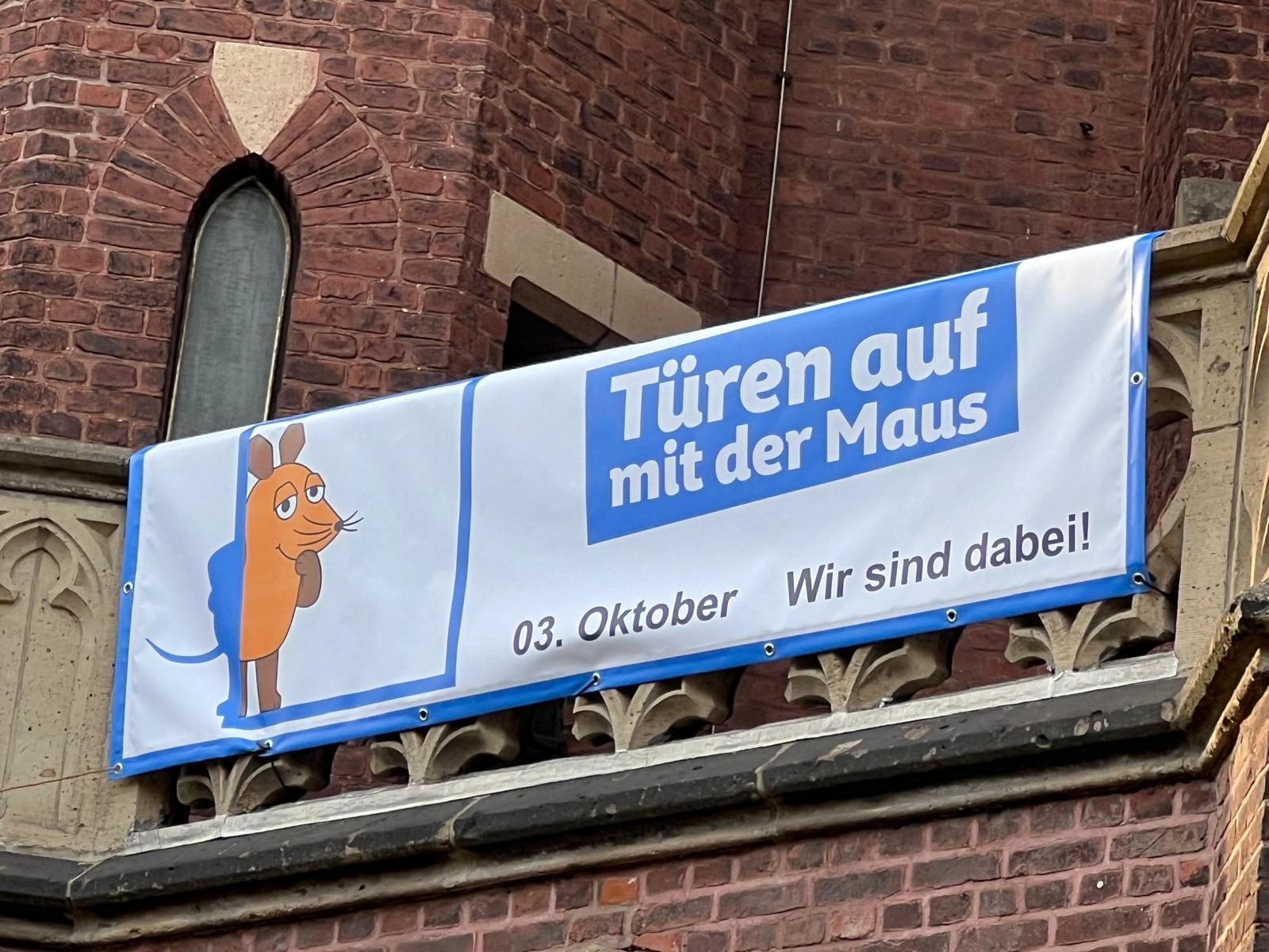 Banner mit der Zeichentrickfigur Maus und dem Text 'Türen auf mit der Maus 03. Oktober Wir sind dabei' an einer Steinbalustrade vor rotem Backsteinmauerwerk.