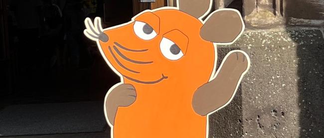 Aufsteller in Form einer orange-braunen Maus mit erhobener Hand vor einer Kirchentür