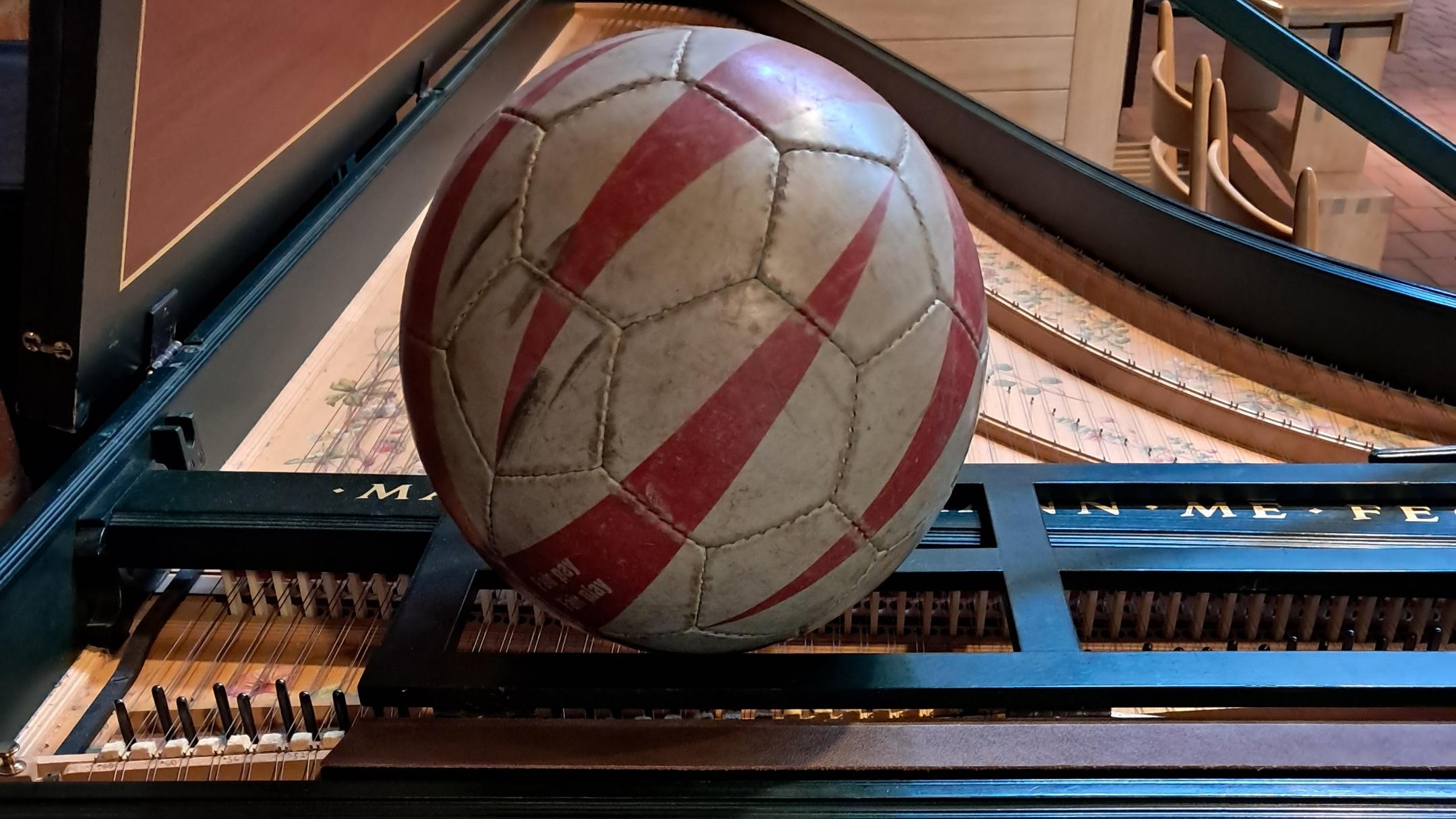 Ein roter weißer Fußball steckt zwischen dem Flügel, erkennbar sind auch die Tasten des Flügels.