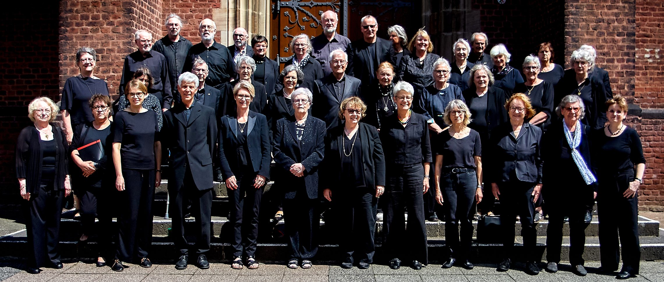 Vor der braunen Kirchentür stehen in drei Reihen viele Frauen und Männer in schwarzer Kleidung.