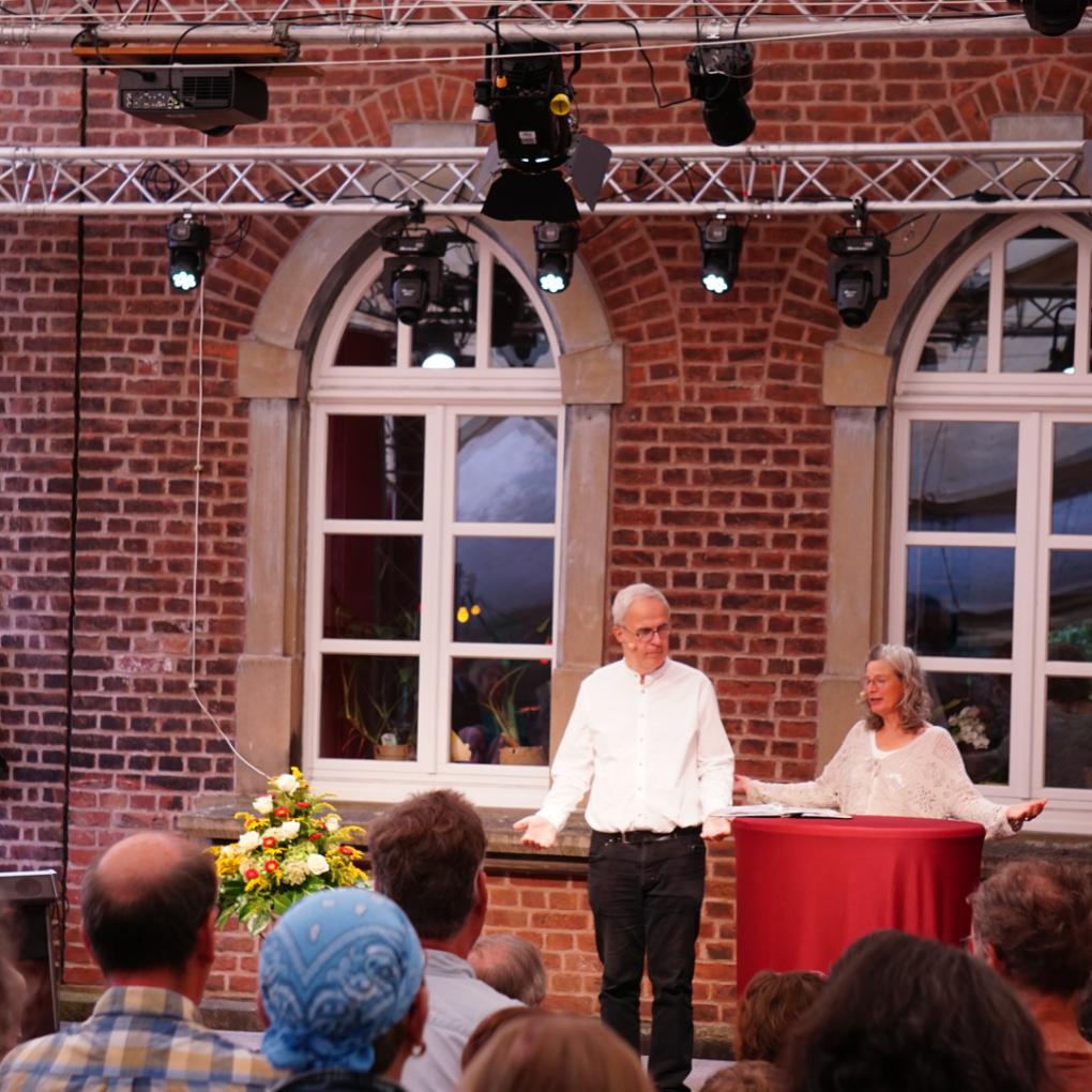 Drei Personen stehen auf einer Bühne vor einem Publikum Open Air vor Backsteinwänden und großen Fenstern.