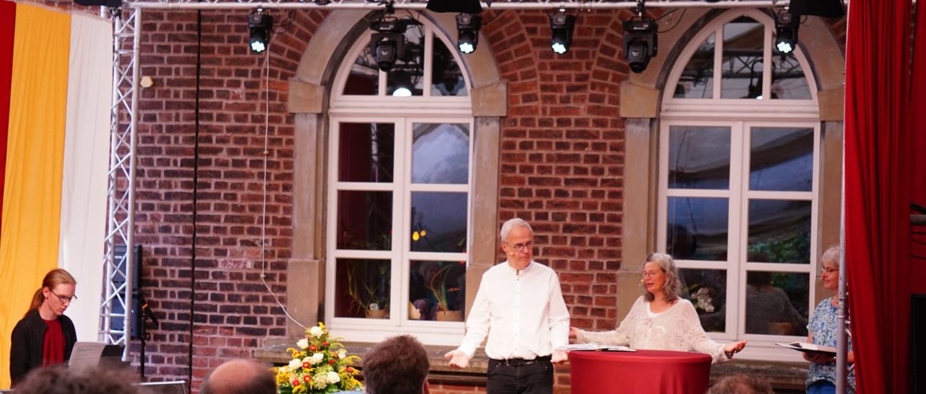 Drei Personen stehen auf einer Bühne vor einem Publikum Open Air vor Backsteinwänden und großen Fenstern.