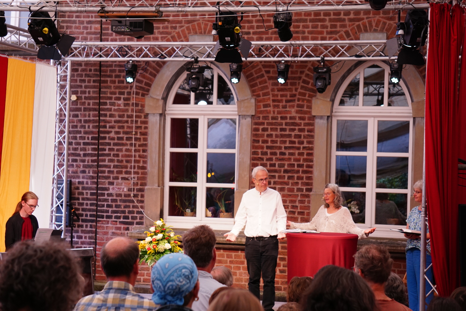 Drei Personen stehen auf einer Bühne vor einem Publikum Open Air vor Backsteinwänden und großen Fenstern.