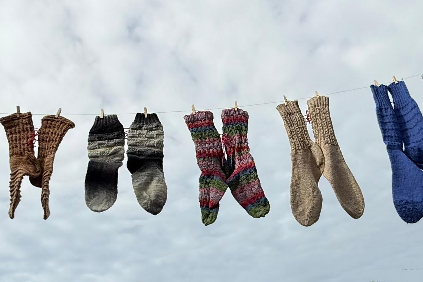 Mehrere bunte gestrickte Socken hängen an einer Wäscheleine vor bewölktem Himmel.