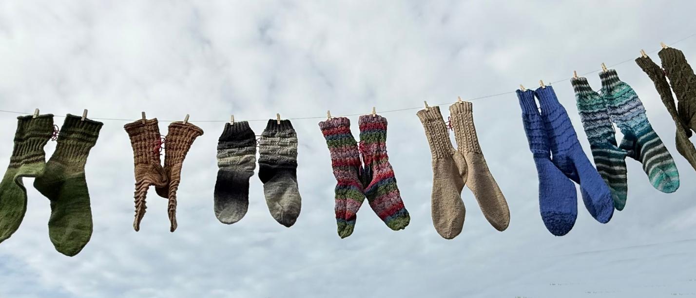 Mehrere bunte gestrickte Socken hängen an einer Wäscheleine vor bewölktem Himmel.