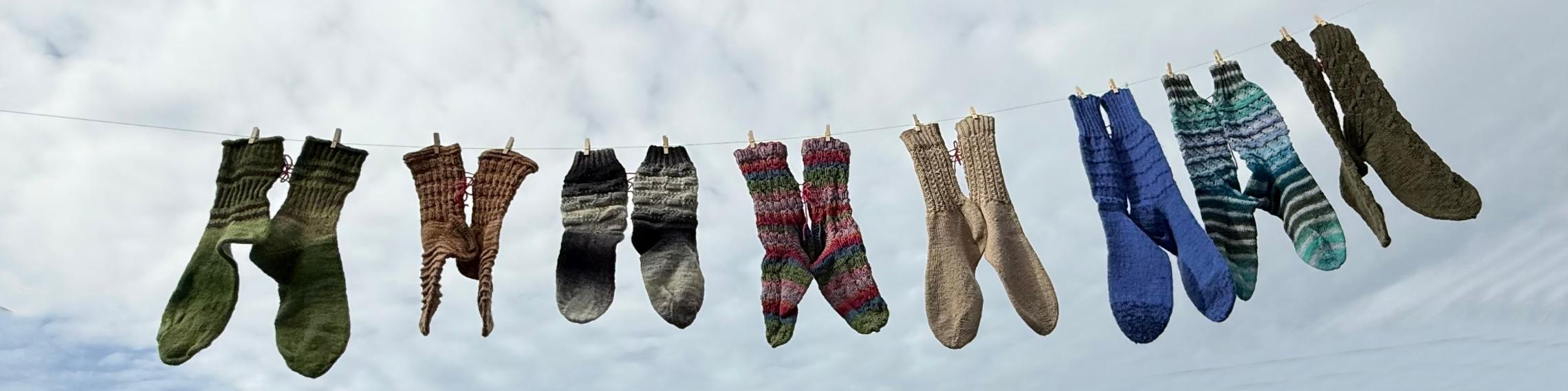 Mehrere bunte gestrickte Socken hängen an einer Wäscheleine vor bewölktem Himmel.