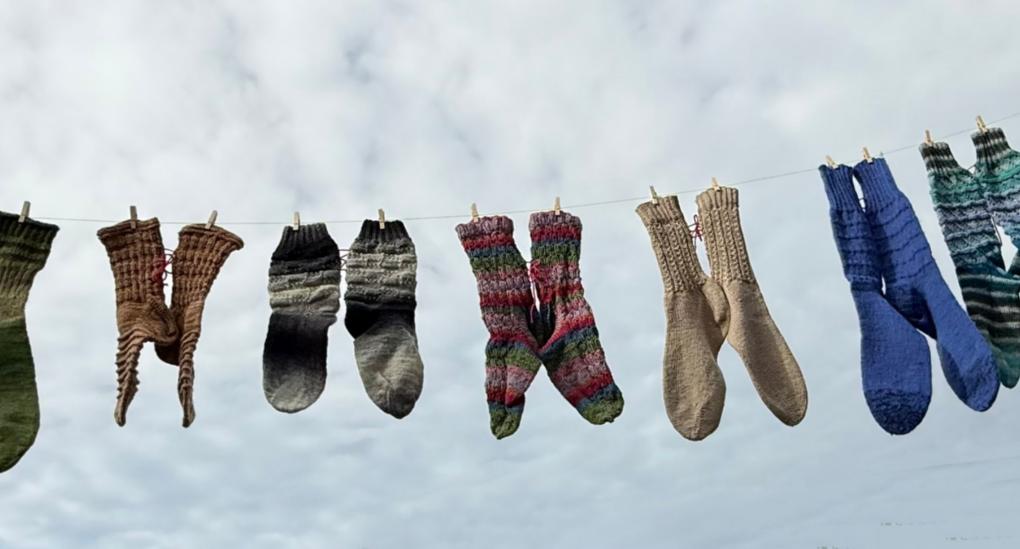 Mehrere bunte gestrickte Socken hängen an einer Wäscheleine vor bewölktem Himmel.