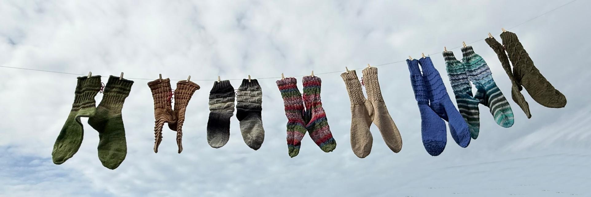 Mehrere bunte gestrickte Socken hängen an einer Wäscheleine vor bewölktem Himmel.