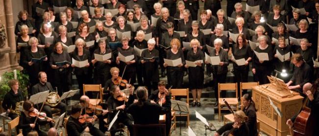 Viele Chormitglieder singen in Formation, davor steht der Dirigent mit Orchester.