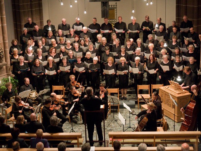 Viele Chormitglieder singen in Formation, davor steht der Dirigent mit Orchester.