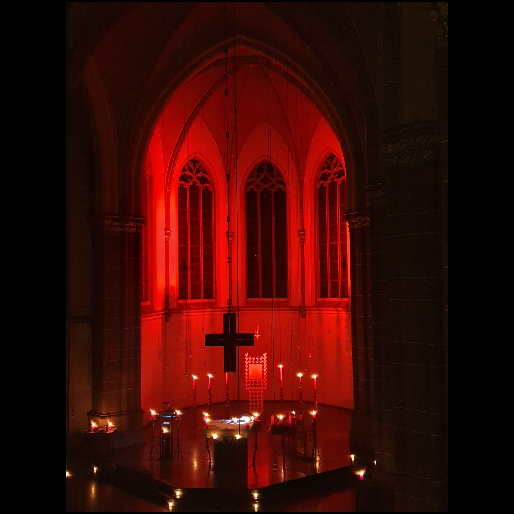 Kirchraum liegt in Dunkelheit nur durch Kerzen, die am Rand der Bänke und vorne im Altar stehen, erleuchtet. Die Apsis wird durch rote Strahler erleuchtet.