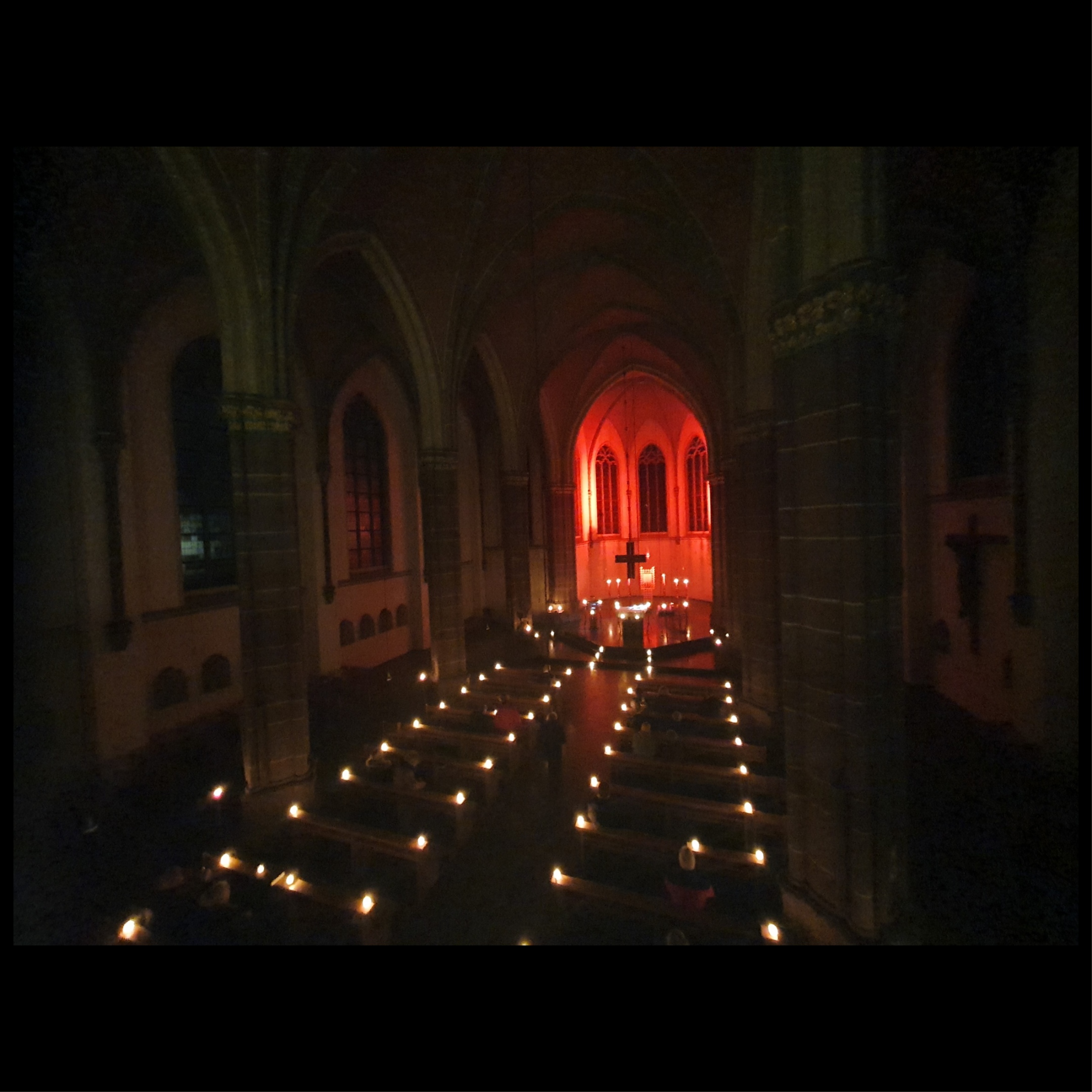 Kirchraum liegt in Dunkelheit nur durch Kerzen, die am Rand der Bänke und vorne im Altar stehen, erleuchtet. Die Apsis wird durch rote Strahler erleuchtet.