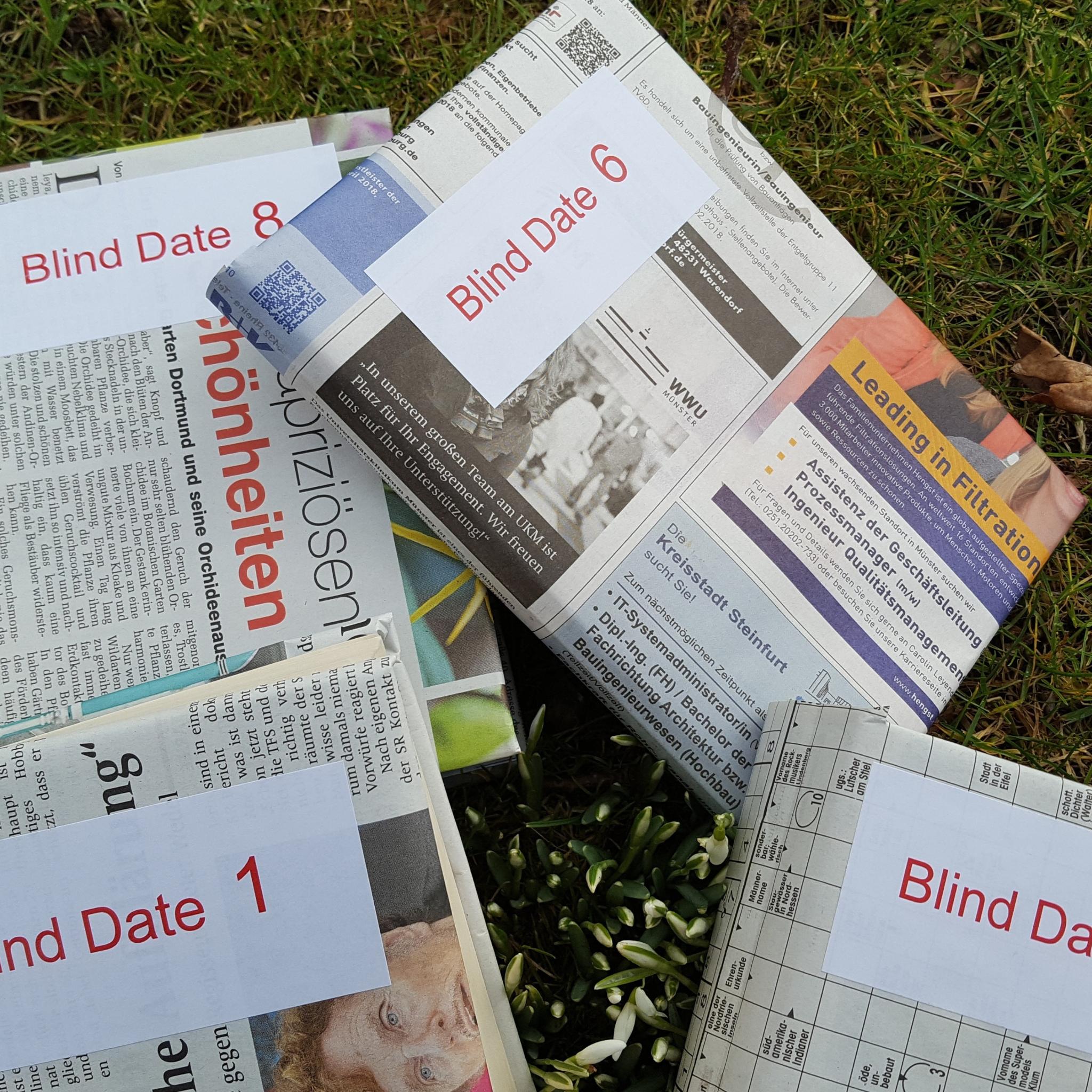 Verschiedene Ausgaben von Zeitungen liegen auf einer grünen Wiese. Auf den Zeitungen ist ein weißer Kasten mit der Aufschrift 'Blind Date' und einer Zahl.
