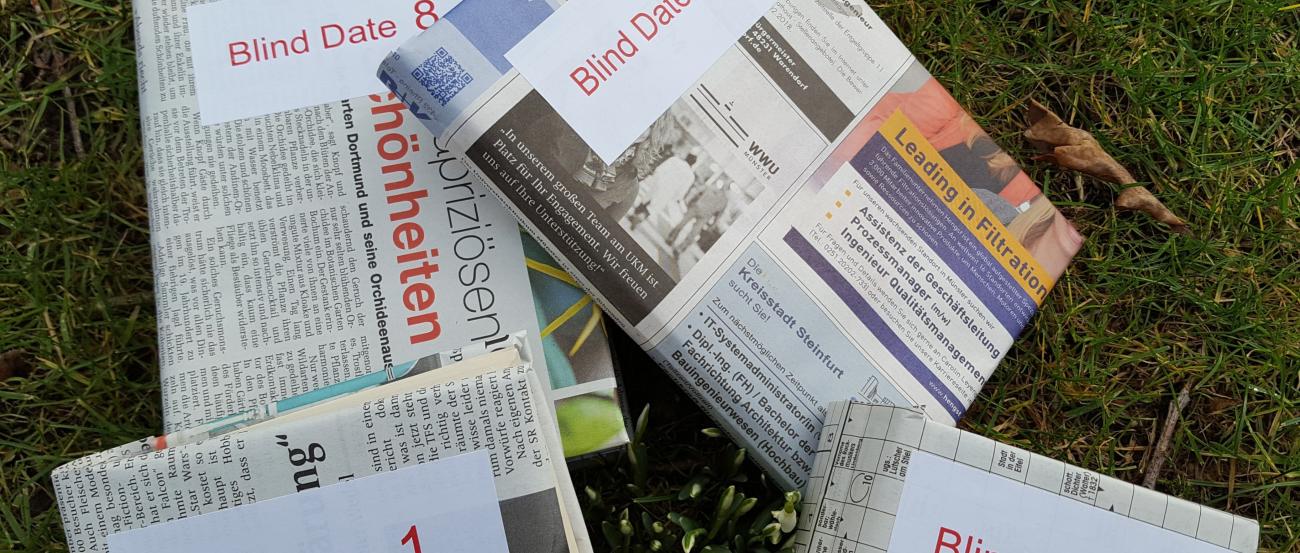 Verschiedene Ausgaben von Zeitungen liegen auf einer grünen Wiese. Auf den Zeitungen ist ein weißer Kasten mit der Aufschrift 'Blind Date' und einer Zahl.