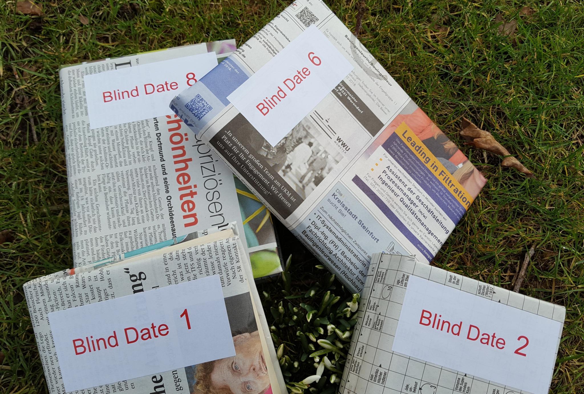 Verschiedene Ausgaben von Zeitungen liegen auf einer grünen Wiese. Auf den Zeitungen ist ein weißer Kasten mit der Aufschrift 'Blind Date' und einer Zahl.