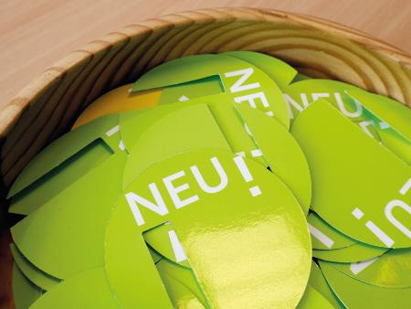 In einer Schale liegen viele grüne Aufkleber mit der weißen Aufschrift 'NEU!'.