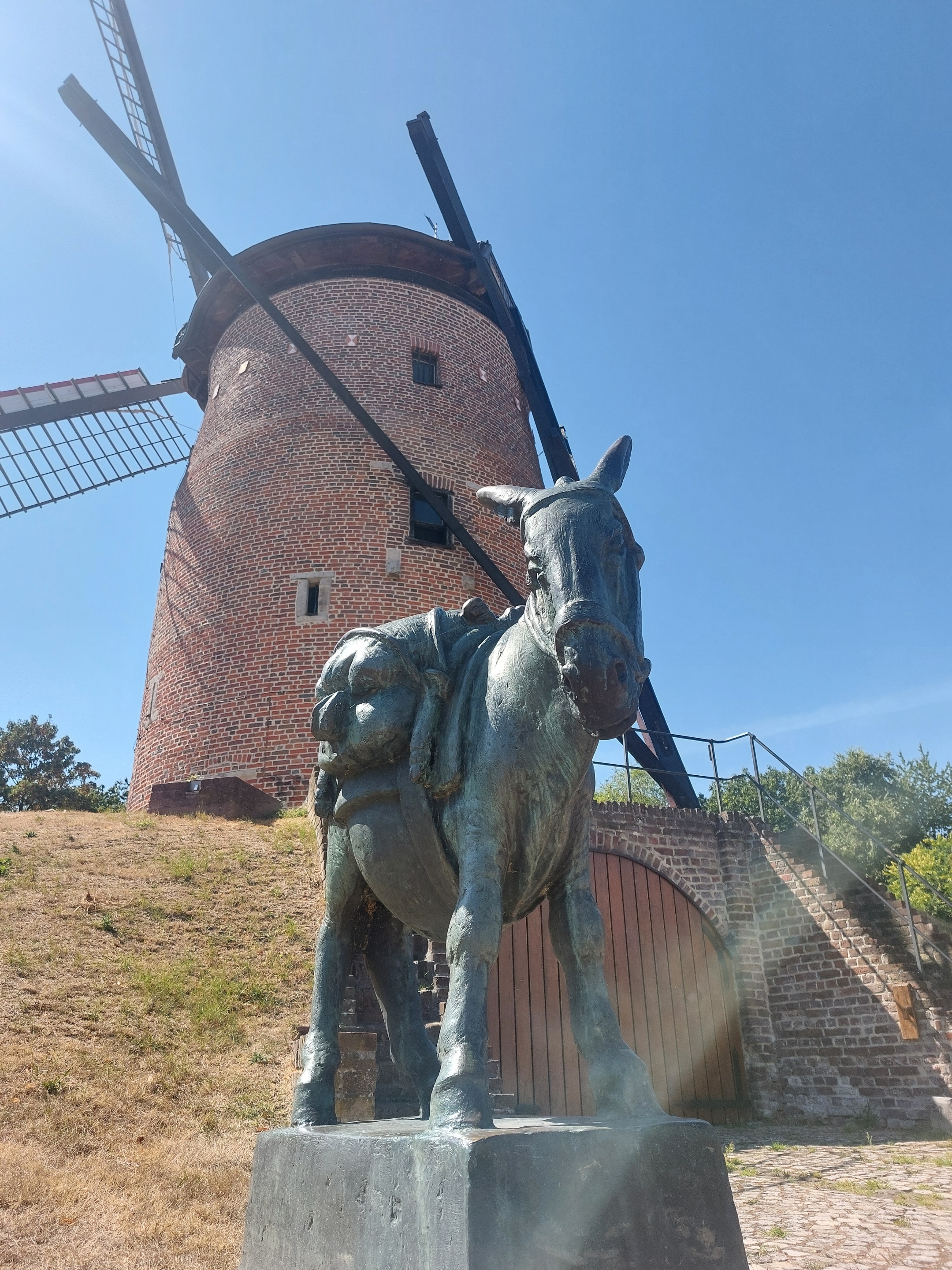 Bronzestatue eines beladenen Esels vor einer historischen Backsteinwindmühle mit Flügeln und Treppe.