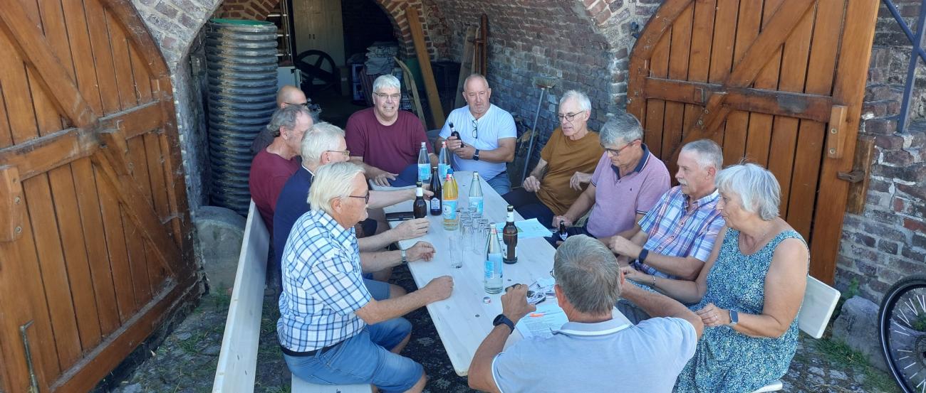Mehrere Menschen sitzen vor einem Tor, die Türen des Tores stehen offen. Die Menschen sitzen auf Bänken.