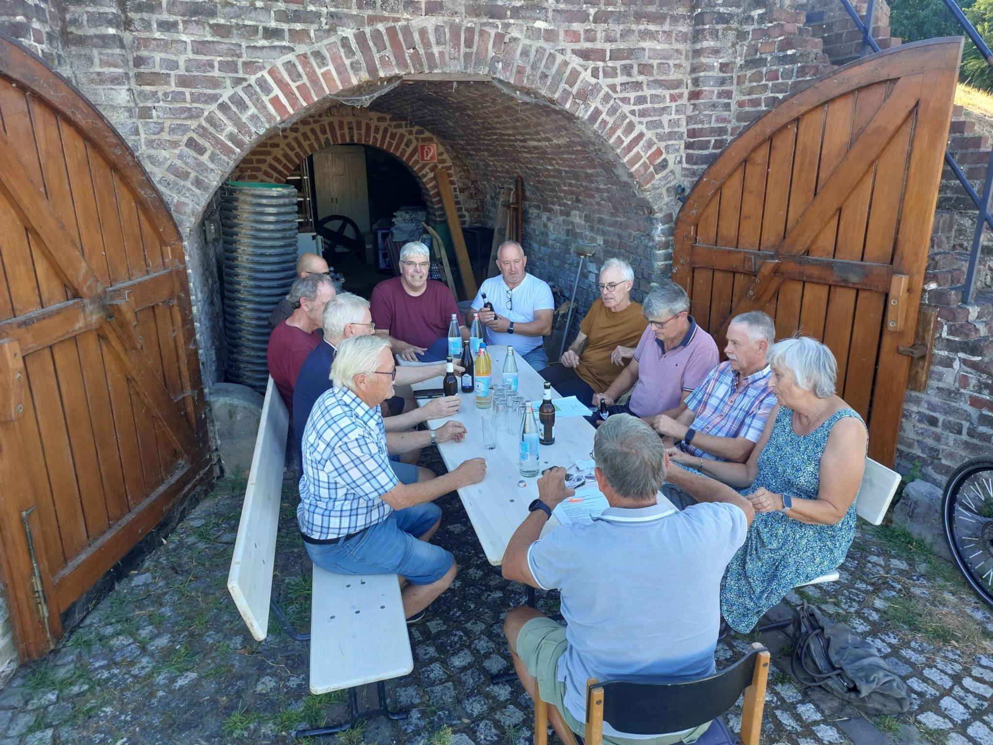 Mehrere Menschen sitzen vor einem Tor, die Türen des Tores stehen offen. Die Menschen sitzen auf Bänken.