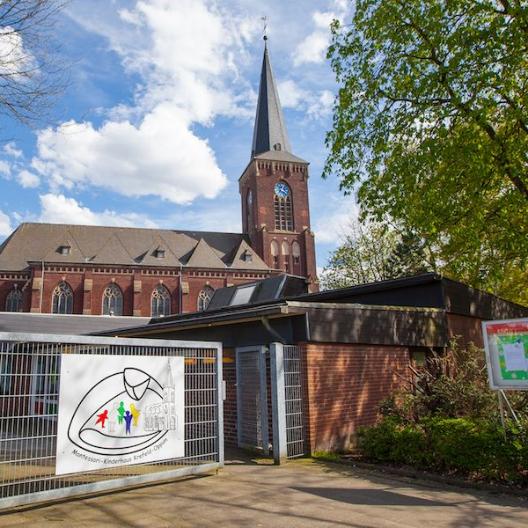 Hellblauer Himmel mit weißen Wolken, im Hintergrund die Kirche Zu den heiligen Schutzengeln, davor das graue Eingangstor zum Kinderhaus mit dem alten Logo