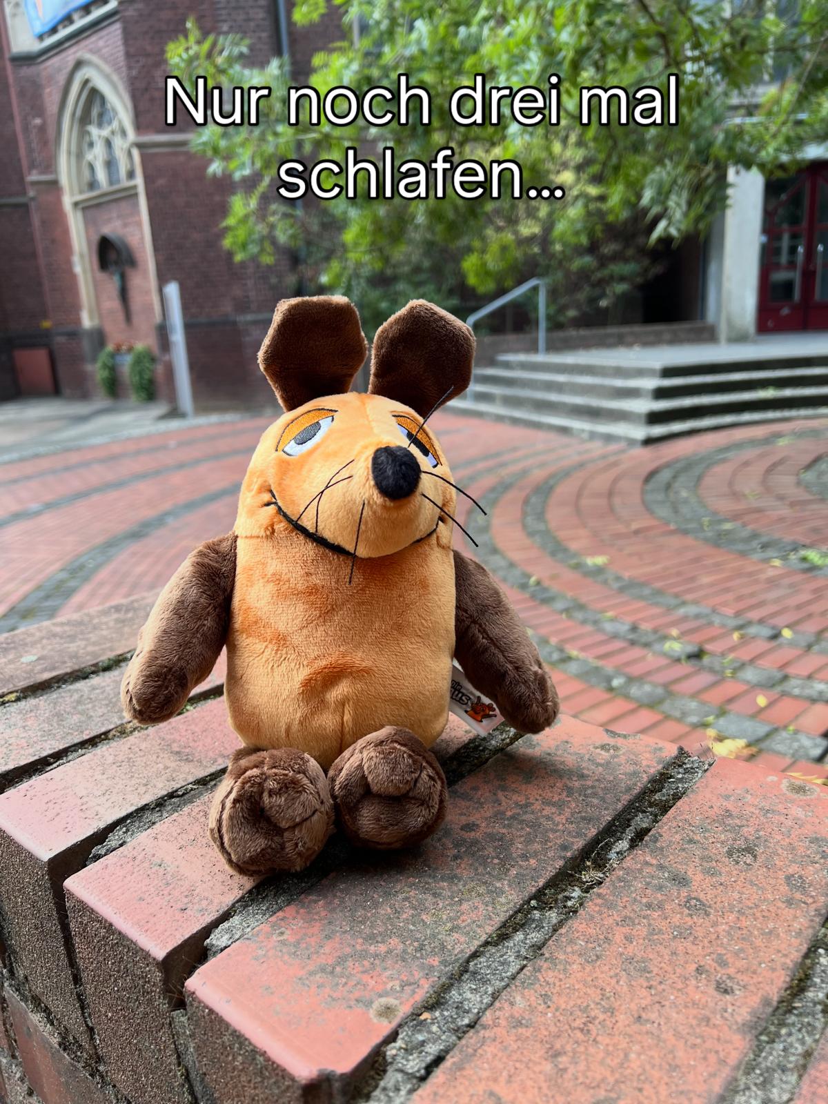 Plüschmaus sitzt auf einer gemusterten Steinbank vor einer Treppe und einem Backsteingebäude, darüber der Schriftzug 'Nur noch drei mal schlafen...'.