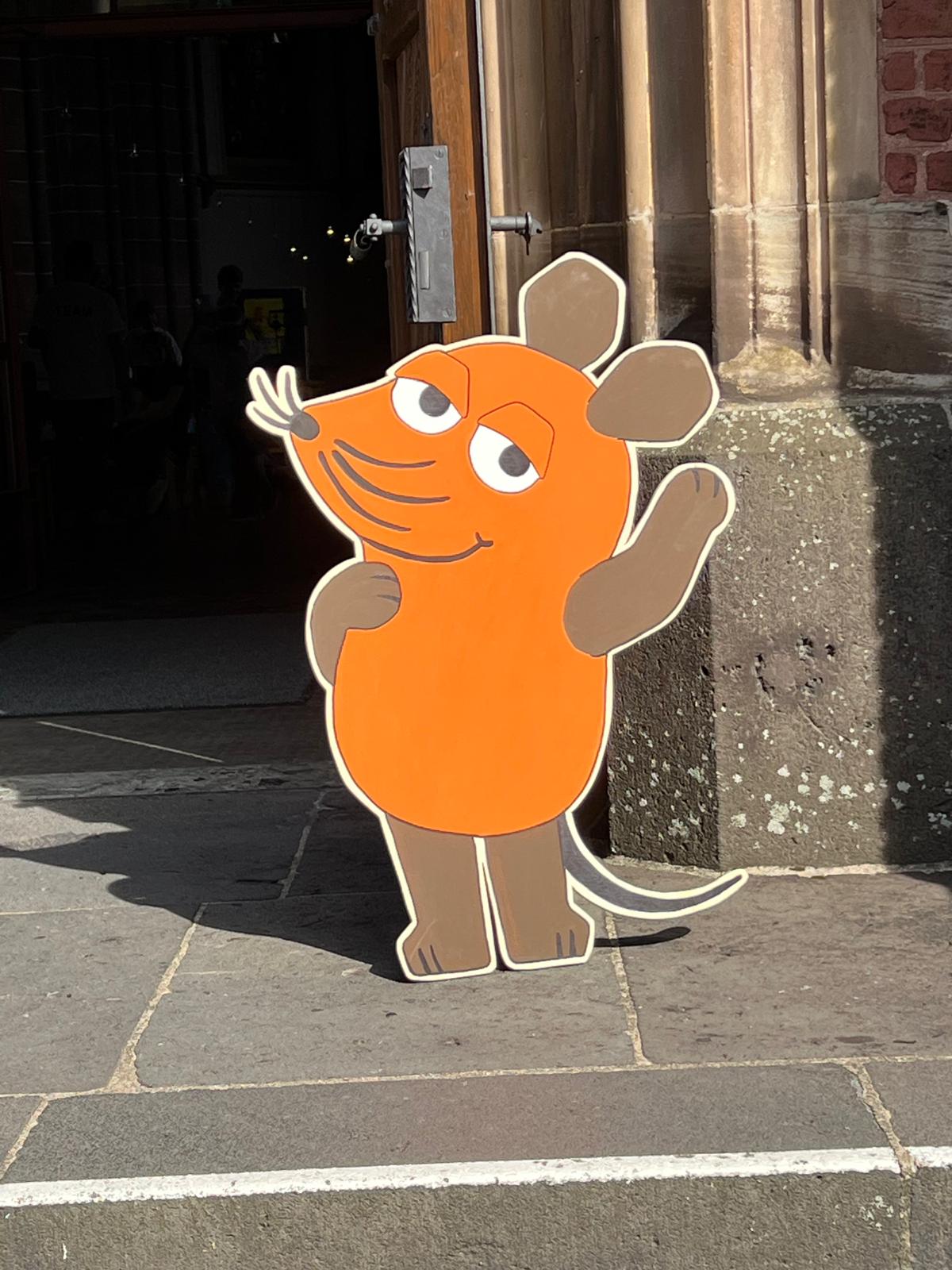 Aufsteller in Form einer orange-braunen Maus mit erhobener Hand vor einer Kirchentür